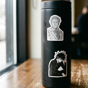 May include: A matte black cylindrical tumbler featuring two distinct anime-style character stickers. The top sticker depicts a man in a suit and tie, while the bottom sticker shows a character with spiky dark hair resting their chin on a hand.