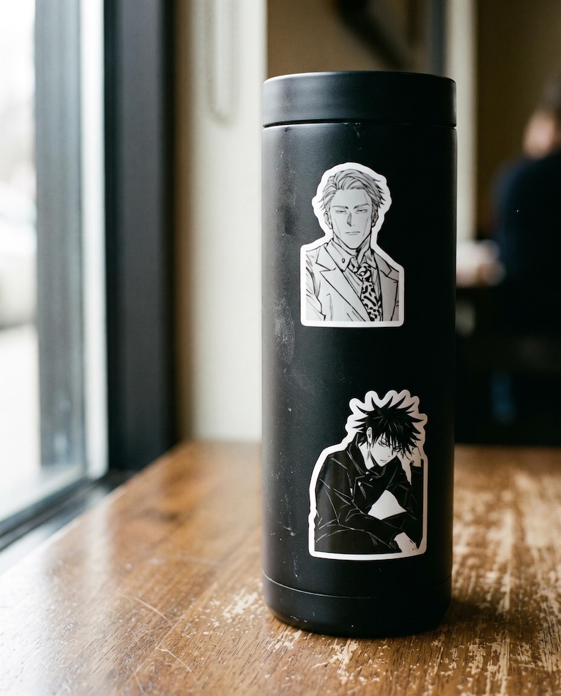 May include: A matte black cylindrical tumbler featuring two distinct anime-style character stickers. The top sticker depicts a man in a suit and tie, while the bottom sticker shows a character with spiky dark hair resting their chin on a hand.