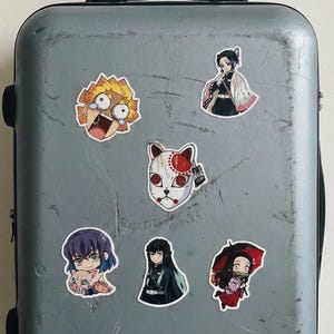May include: A selection of anime character stickers, including a character with a fox mask and a character with spiky yellow hair. The stickers are attached to a grey suitcase, showcasing a variety of designs and expressions.