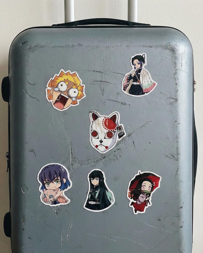 May include: A selection of anime character stickers, including a character with a fox mask and a character with spiky yellow hair. The stickers are attached to a grey suitcase, showcasing a variety of designs and expressions.