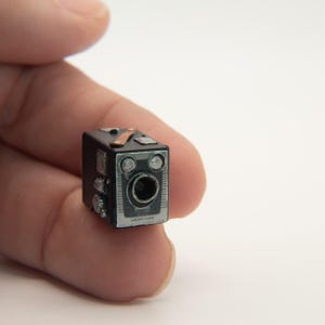 May include: Miniature black and silver vintage camera with a brown strap detail. The camera has a detailed front with a lens and two circular accents. The camera is held in a person's hand.