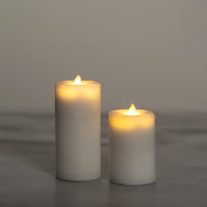 May include: Two white, battery-operated pillar candles with a warm, flickering LED light. The candles are different sizes, with one taller than the other. The candles are on a light grey surface.