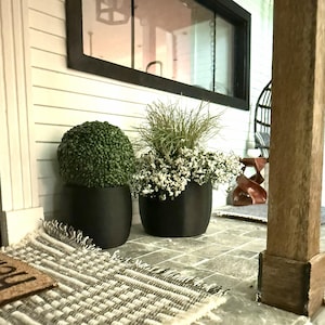 May include: A miniature outdoor scene featuring two black planters with greenery. One planter holds a round, green topiary, and the other has white flowers and tall grass. A woven rug and a window are in the background.