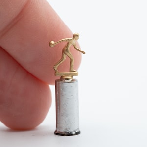 May include: A miniature gold-colored figurine of a person in a bowling stance, mounted on a small, cylindrical, silver-toned metal base. The figure is depicted in a dynamic motion, holding a bowling ball, and is presented as a tiny trophy.
