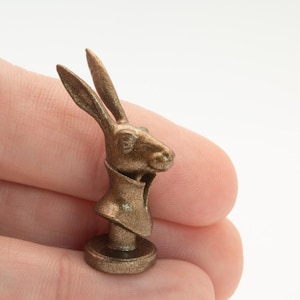 Könnte beinhalten: Eine kleine, bronzefarbene Metallfigur, die den Kopf und Hals eines Hasen darstellt. Der Hase ist im Profil zu sehen, trägt ein Kleidungsstück mit hohem Kragen und ist auf einem runden Sockel montiert. Die Oberfläche hat ein strukturiertes Finish.