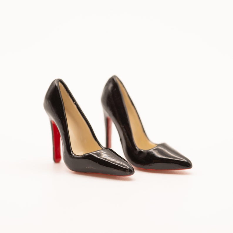 Puede incluir: Un par de zapatos de tac&oacute;n negros con punta y suelas rojas. Los zapatos brillantes est&aacute;n sobre un fondo blanco. Los tacones miden aproximadamente 10 cm de alto.
