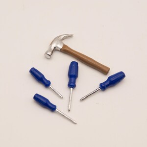 May include: A miniature claw hammer with a silver-toned metal head and a brown handle, accompanied by four small screwdrivers featuring bright blue plastic handles and thin silver-toned metal shafts, arranged together on a plain surface.
