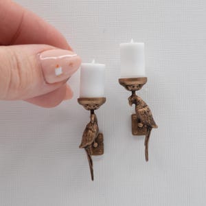 May include: Two miniature wall-mounted candle holders crafted from a bronze-toned metal, each featuring a detailed parrot figure supporting a small, cylindrical white candle. The parrot designs are textured and include a square mounting plate.