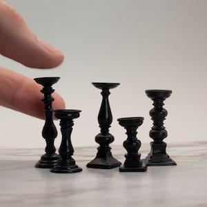 May include: Five miniature black candle holders of varying heights and designs. The candle holders have a classic, ornate appearance, with a small cup at the top to hold a candle. The bases are square or round.