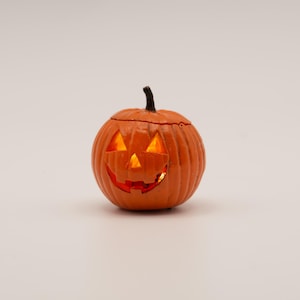 May include: A small, round, orange jack-o'-lantern figurine with a dark brown stem. The pumpkin features carved triangular eyes and a jagged mouth, illuminated from within by a warm, glowing light that shines through the cut-out facial features.