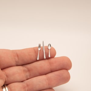 May include: Miniature silver fork, knife, and spoon set. The tiny silverware is arranged on a person's finger, showcasing the small size and detail of the cutlery. The set is likely for a dollhouse or miniature display.