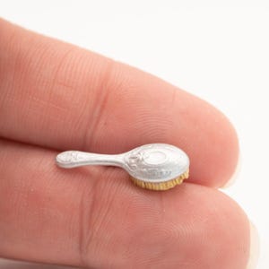 May include: A miniature silver-colored hairbrush with a decorative floral design on the handle and brush head. The bristles are a light tan color. The brush is small, likely for a doll or display.