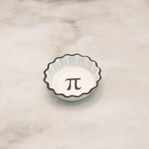 May include: A small, white, scalloped-edge dish with a black rim. The Greek letter pi, π, is centered in black. The dish is on a light gray marble surface.