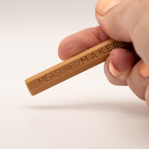 May include: A rectangular, light brown object with the text "MEASUREDMAKER" engraved on its surface. The object is held by a person with fair skin and orange nail polish. The background is a soft white.