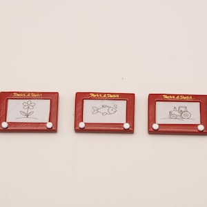 May include: Three miniature red Etch-A-Sketch toys, each with a different drawing: a flower, a fish, and a tractor. The toys have white knobs and the words "Sketch A Sketch" printed at the top.