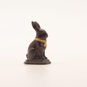 May include: A dark brown chocolate bunny figurine with a gold-colored collar. The bunny is sitting upright on a small, rectangular base. The figurine is set against a plain white background.