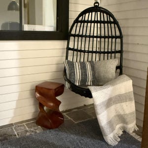 May include: A black wicker hanging chair with two pillows and a striped blanket. A wooden side table is to the left. The chair is hanging on a porch with white siding and a blue rug.