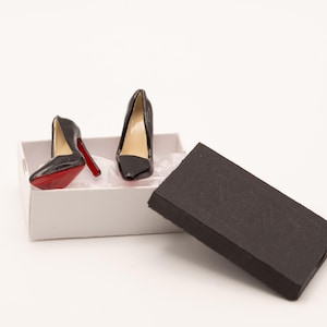 May include: Miniature black high heels with red soles are displayed in a white box with a black lid. The heels have a glossy finish and a classic pointed-toe design. The box is open, revealing the shoes.