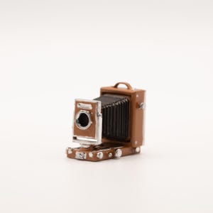 May include: A miniature brown and silver vintage camera with a bellows design. The camera has a handle on top and a lens with a silver frame. The camera is a detailed replica of a classic camera.