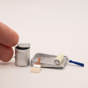 May include: A miniature silver paint can with a lid, a small paint tray, a paintbrush with a brown handle and white bristles, and a paint roller with a blue handle. These tiny painting tools are arranged together on a plain white surface.