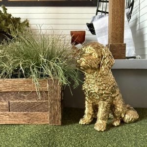 Könnte beinhalten: Eine goldfarbene Hundefigur mit strukturierter Oberfläche, die auf einer grünen Oberfläche sitzt. Der Hund hat lockiges Fell und befindet sich in der Nähe eines hölzernen Pflanzkastens mit grünem Gras. Die Szene spielt auf einer Veranda.