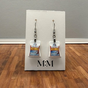 May include: A pair of novelty earrings featuring a miniature ice melt bag design. Each white bag has the text "STAY SALTY ICE MELT" printed on it, with a blue and white graphic. The earrings hang from silver-colored hooks.