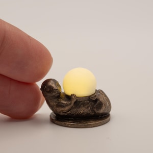 May include: A bronze-coloured figurine of a small animal holding a glowing yellow orb. The figurine is resting on a circular base. The orb emits a soft, warm light. The figurine is held by a person's finger.