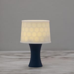 Puede incluir: Una pequeña lámpara de mesa azul marino con una pantalla blanca. La pantalla tiene un patrón de recortes circulares espaciados uniformemente que emiten un brillo cálido. La base de la lámpara tiene forma de reloj de arena curvado.