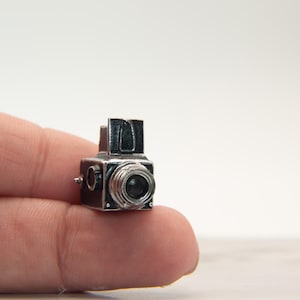 May include: A miniature, vintage-style camera with a black body and silver-colored lens and details. The small camera is held between two fingers. The camera has a rectangular shape with a lens and a viewfinder.