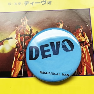 May include: A light blue button with the word "DEVO" in large black letters. Below the word "DEVO" is the text "MECHANICAL MAN" in smaller black letters. The button is on a yellow surface with a band photo in the background.