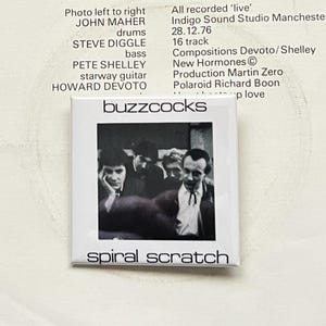May include: A square button featuring the band name "buzzcocks" above a black and white photo of the band members. The text "spiral scratch" is printed below the photo. The button is likely a collectible item.