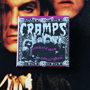 May include: Square button featuring the band The Cramps with the words "GARBAGEMAN" and "DRUGTRAIN" below a band photo. The button has a purple border with skulls and the band's name in white.