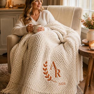 May include: A cream-colored chunky knit throw blanket featuring a textured bobble pattern and long fringe edges. The blanket is embroidered with a rust-colored laurel branch design, the initials AR, and the year 2026 in a serif font.