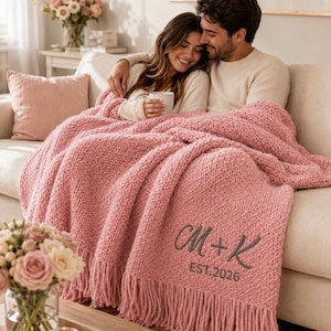 Puede incluir: Una manta de punto grueso en color rosa empolvado con un borde de flecos en la parte inferior. La manta está bordada con las letras M + K en una fuente cursiva de color gris oscuro, seguidas del texto EST. 2026 en una fuente sans-serif simple.