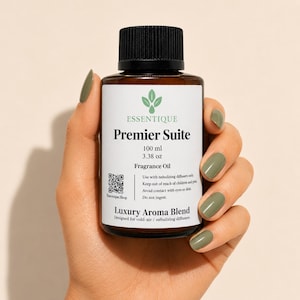 以下が含まれることがあります： 黒いキャップが付いた、Premier Suiteフレグランスオイルの茶色のガラス瓶、100 ml。ラベルにはブランド名、製品名、使用方法が記載されています。オリーブグリーンのマニキュアを塗った手でボトルを持っています。