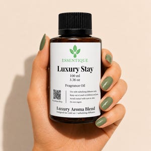 May include: A brown glass bottle with a black cap, held by a hand with green nail polish. The label reads "ESSENTIQUE Luxury Stay Fragrance Oil" with a volume of 3.38 oz. The product is designed for cold-air/nebulizing diffusers.