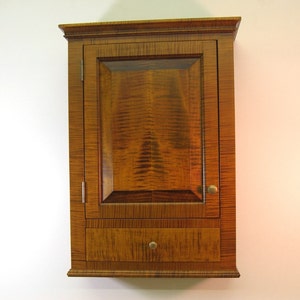 May include: A wall-mounted wooden cabinet with a single door and a drawer. The cabinet is made of a figured wood with a warm, brown tone. The door has a recessed panel and a small, round knob. The drawer also has a round knob.