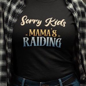 Könnte beinhalten: Schwarzes T-Shirt mit dem Text "Sorry Kids Mama's Raiding" in einer stilisierten Schriftart. Der Text ist in einer Mischung aus Farben, darunter Creme, Gold und Blau. Das Shirt wird mit einem schwarz-weiß karierten Hemd getragen.