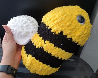 May include: A large, plush, handmade bee-shaped cushion. The bee features a yellow body with black stripes, a white fluffy tail, and a large, black eye. The cushion is made of soft, textured yarn.