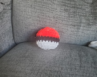 May include: A handmade crochet Pokeball. The toy is red on top, white in the middle, and has a black band. It is made of soft yarn and sits on a grey, textured surface.