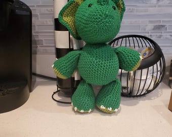 May include: A green crochet dinosaur toy with yellow detailing on the wings and feet. The dinosaur has a rounded body, small limbs, and a large head with horns. A charming, handmade item.