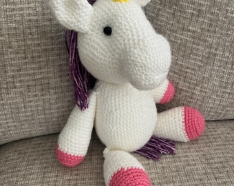 May include: A white crochet unicorn toy with a yellow horn, purple mane, and pink hooves. The soft toy is resting on a grey surface. The unicorn has black eyes and is made of soft yarn.