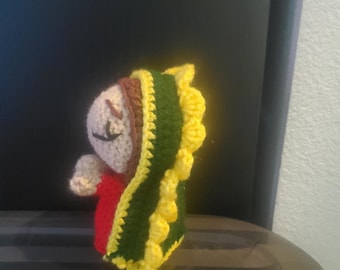 May include: Handmade crocheted doll with a red dress, beige face, and a green and yellow shawl. The doll has closed eyes and small arms. The shawl has yellow, scalloped edges. A unique, handcrafted item.
