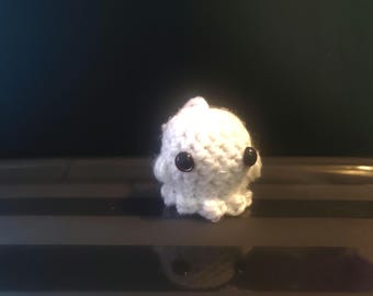 May include: A small, white crocheted octopus with black button eyes. The handmade amigurumi figure has a rounded body and eight small tentacles. The octopus is sitting on a dark surface, with a dark background.