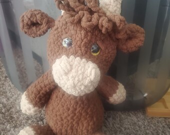 May include: A handmade crochet cow plushie in shades of brown and cream. The plush toy features a brown body, head, and ears, with cream-coloured accents on the face, hooves, and horns. The cow has large, shiny eyes.