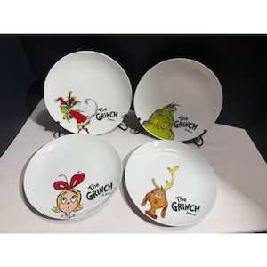 Puede incluir: Un juego de cuatro platos de cerámica blanca con ilustraciones coloridas de los personajes de El Grinch. Cada plato muestra un gráfico único que incluye al Grinch con traje de Santa, su rostro, Cindy Lou Who y Max el perro con astas.