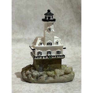 May include: A miniature lighthouse figurine. The lighthouse is white with a black top and black shutters. It sits atop a stone base with a door and is surrounded by a rocky, textured foundation. The overall design is detailed and decorative.