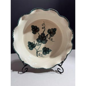 May include: A round ceramic pie dish with a cream-colored interior and a scalloped edge rimmed in dark green. The center of the dish features a hand-painted botanical design of dark green leaves and swirling vines on a smooth, glazed surface.