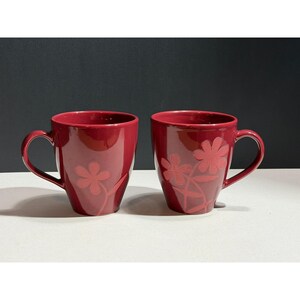 May include: Two red ceramic mugs with a floral design. The mugs have a curved handle and a glossy finish. The floral design is a lighter shade of red and is etched into the surface of the mugs. The mugs are on a white surface.