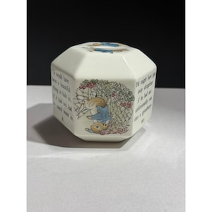 May include: An off-white, octagonal ceramic container featuring a colorful illustration of a brown rabbit wearing a blue jacket caught in a green gooseberry net. Black printed text surrounds the image, detailing the scene from a storybook.